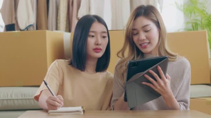 年轻的亚洲夫妇妇女在出售衣服后占收入和支出。几个女人坐在一张小桌子旁，用平板电脑在家做账。