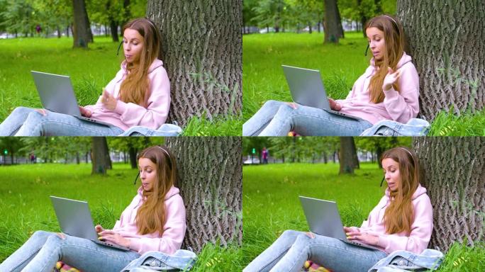 微笑的女人坐在城市公园里，戴着笔记本电脑和耳机进行在线对话。