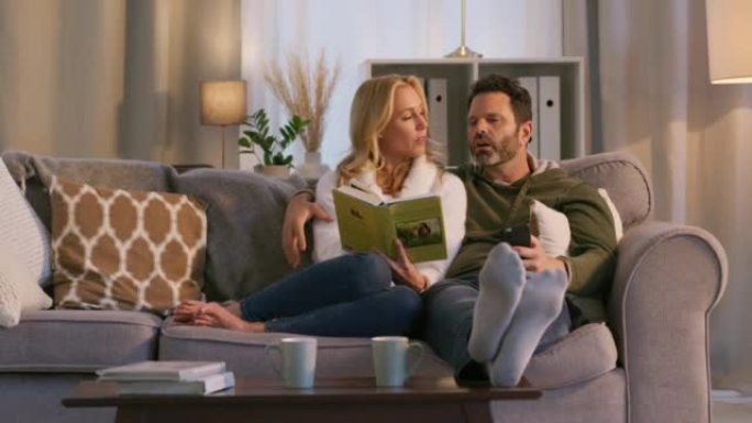 放松的女人向丈夫解释她的书。已婚夫妇一起在沙发上放松，阅读和使用手机。快乐的丈夫拥抱，用智能手机拥抱