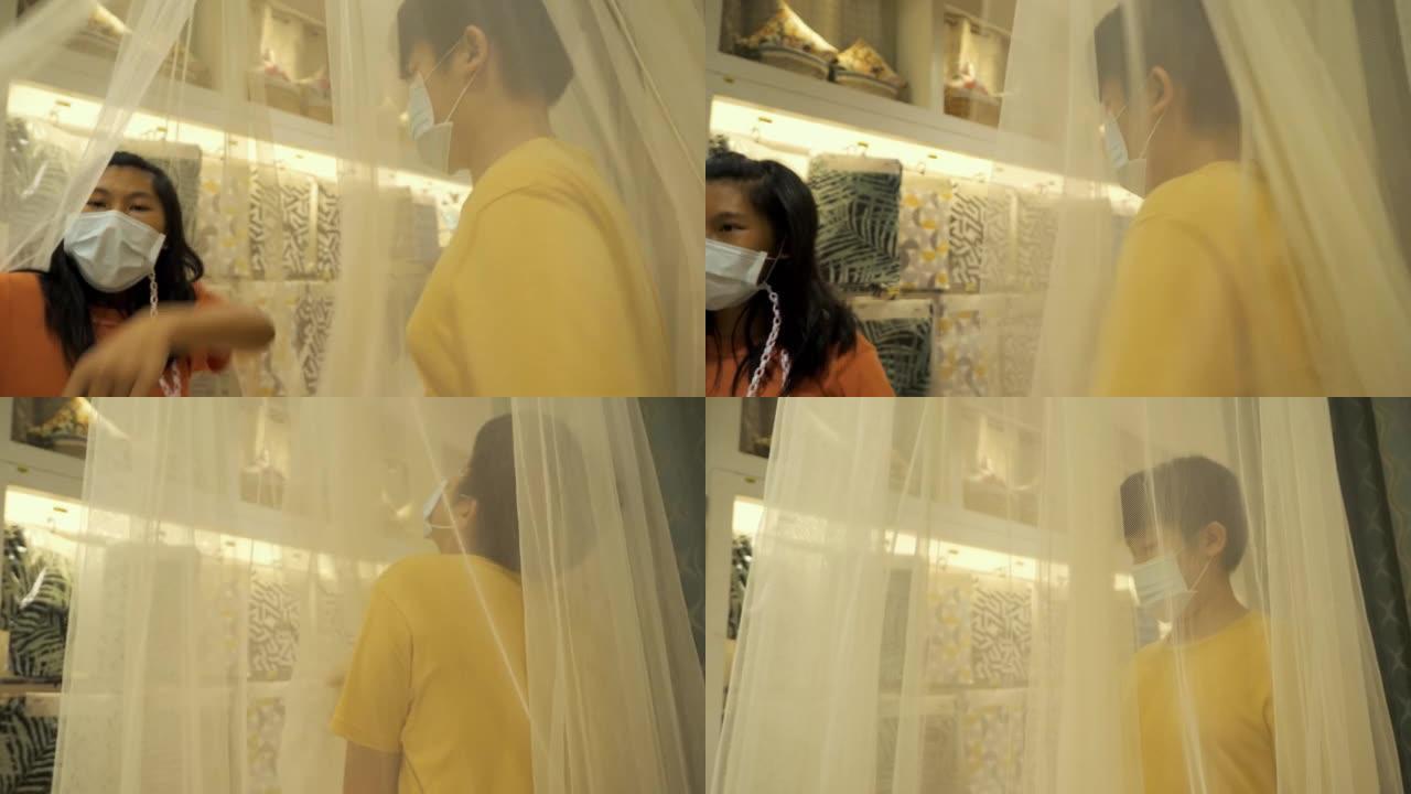 亚洲儿童戴着口罩在家具店玩窗帘，以了解购物生活方式的概念。
