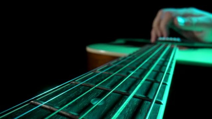 男子音乐家在黑底绿光下弹奏木制原声吉他。手指触摸琴弦，使它们振动并演奏音乐旋律。吉他演奏和弦特写