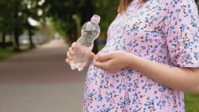 一名孕妇穿着带有花卉图案的夏装站在公园里，喝着富含微量元素的纯净泉水。一名孕妇的腹部和一瓶水的特写镜