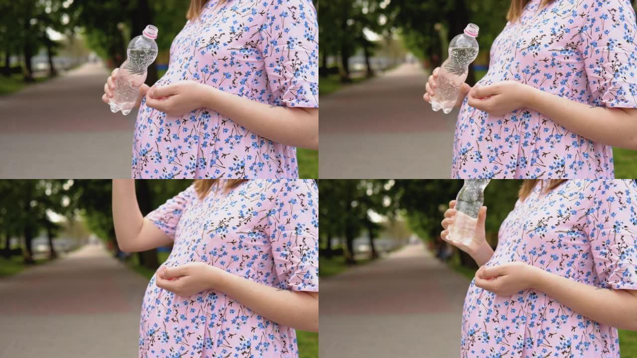 一名孕妇穿着带有花卉图案的夏装站在公园里，喝着富含微量元素的纯净泉水。一名孕妇的腹部和一瓶水的特写镜