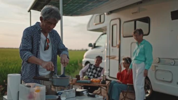 亚洲华人老人在周末休闲活动中为停在稻田的露营车旁边的高级朋友做饭