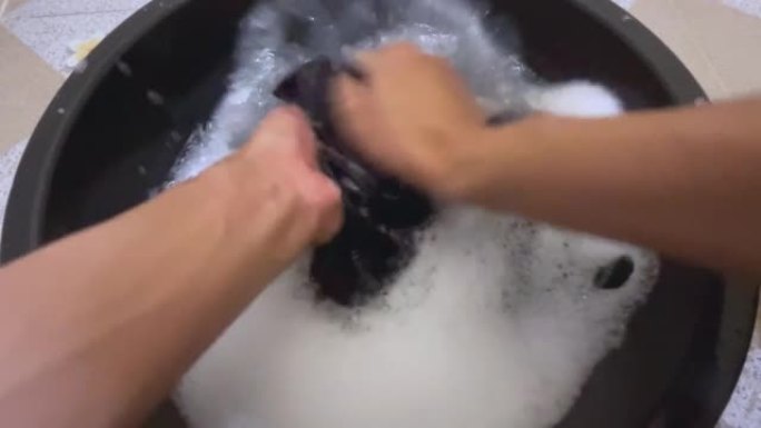 在第一人称视角中，男人的手在塑料盆里洗一件脏衬衫。