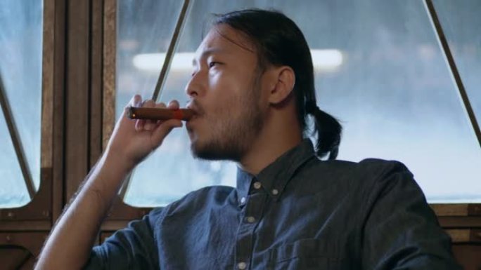 一个年轻的亚洲男子坐着抽雪茄，享受雪茄。一名戴着眼镜和发带的男子坐在酒店的沙发上抽着雪茄吧。