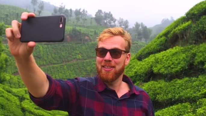 戴着墨镜的红发姜大胡子男人通过智能手机视频聊天，向印度munnar kerala茶园问好