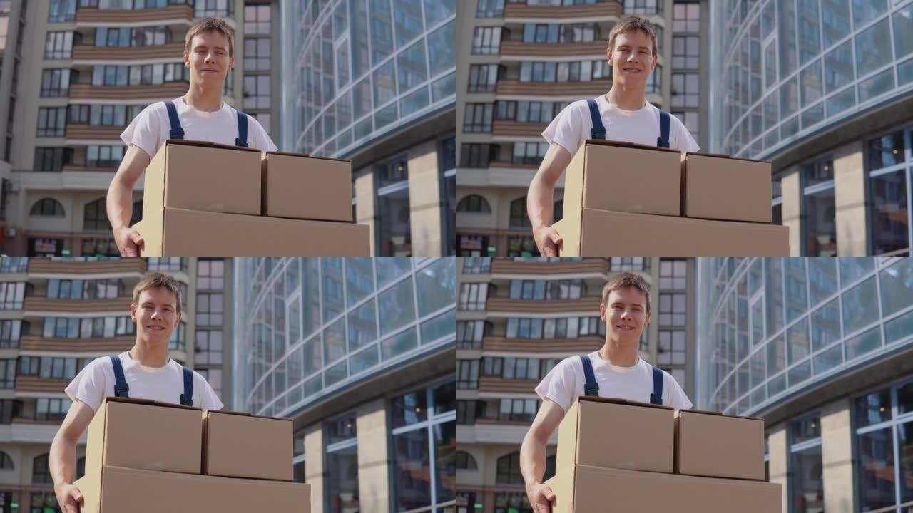 穿着白色t恤和蓝色工作服的送货员在现代高层建筑的背景下拿着三个盒子。快递