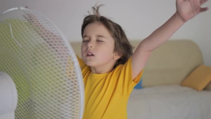夏天炎热的天气，孩子在家享受电风扇带来的凉风。孩子站在客厅家里的风扇热天。夏天炎热的孩子享受新鲜的凉