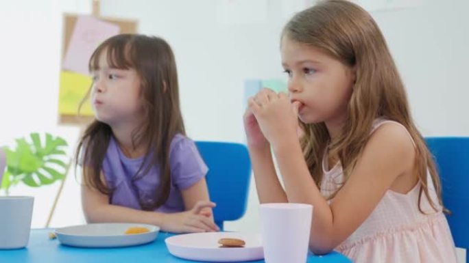 两个年轻的高加索儿童学生一起在桌子上吃午饭。在幼儿园托儿所的教室里，小学龄前女孩在课间休息时感到快乐