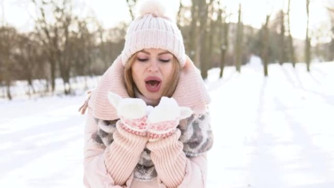 年轻女子穿着柔软的粉红色羽绒服，白帽子，连指手套和围巾在雪地上吹