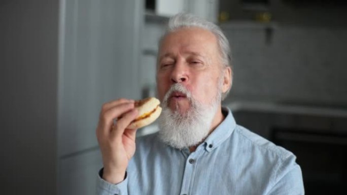 快乐的成年老人站在厨房里闭着眼睛吃美味的芝士汉堡的肖像。