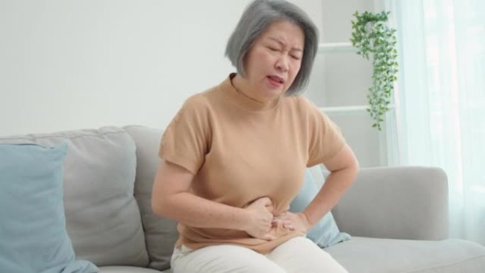 亚洲老年妇女坐在沙发上，双手抓着肚子，因严重腹痛、腹泻、胃炎、肠炎独自在客厅。人们的健康问题观念。