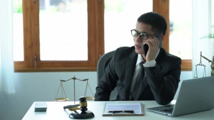 一名亚洲男性律师正在办公室里用智能手机向客户咨询，他在法庭的桌子上放着木锤，在女神天平上放着笔记本电