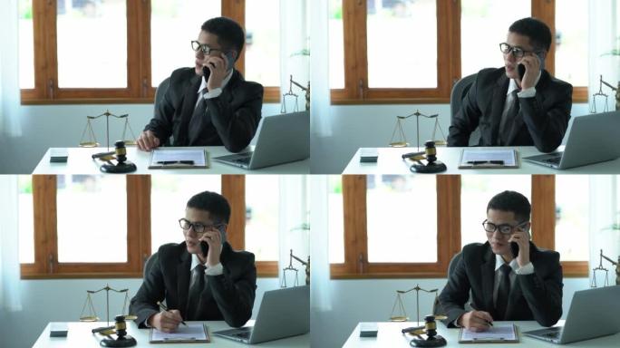 一名亚洲男性律师正在办公室里用智能手机向客户咨询，他在法庭的桌子上放着木锤，在女神天平上放着笔记本电