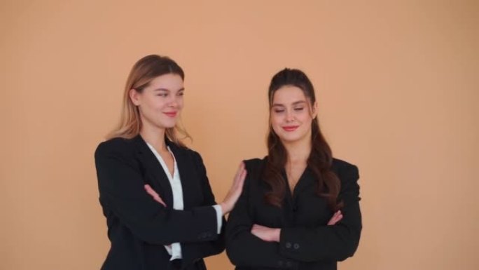 两名年轻的女商人穿着黑色正装站立工作室米色背景看起来相机双手交叉在胸前，然后金发开玩笑地推伙伴。商业