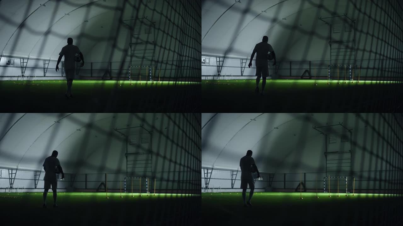 足球运动员在室内训练足球场中，手拿球走路，通过大门上的网观看