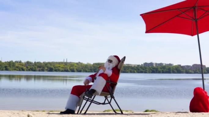 戴着墨镜的圣诞老人躺在湖边海滩的日光浴躺椅上抽雪茄