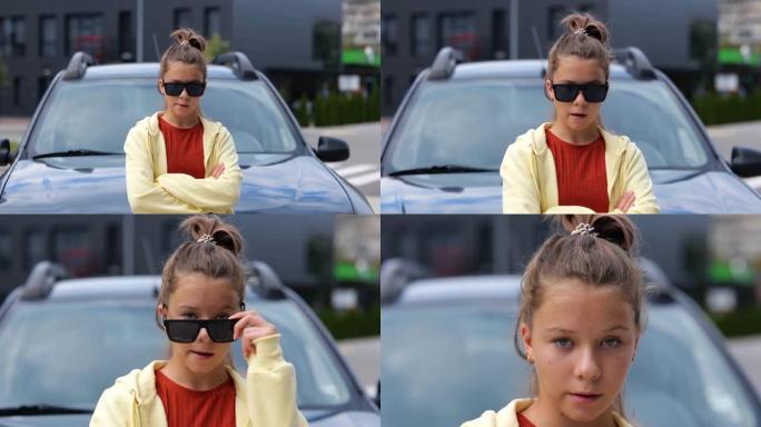 穿着黑色太阳镜的年轻少女站在街上的车前看着相机嚼口香糖。特写。时尚自命不凡的青少年对着镜头微笑。