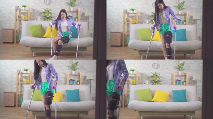 肖像年轻亚洲妇女受伤后膝盖弯曲矫形器膝盖支架用拐杖从沙发上站起来