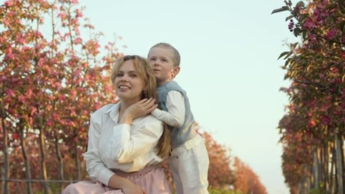妈妈和儿子在花园里的街道上拥抱着粉红色的小树。一个小绅士。父母和孩子之间的爱。继承人，继承