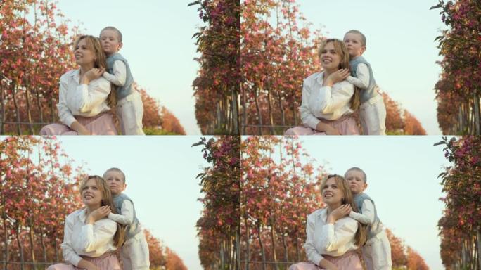 妈妈和儿子在花园里的街道上拥抱着粉红色的小树。一个小绅士。父母和孩子之间的爱。继承人，继承