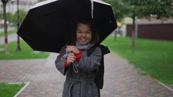 微笑开朗的小女人旋转雨伞的肖像挂在肩膀上的雨衣配件看着相机。阴天，快乐快乐的高加索小人在户外的城市街