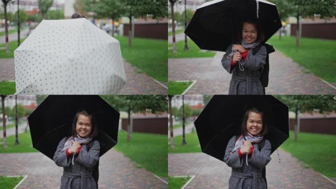 微笑开朗的小女人旋转雨伞的肖像挂在肩膀上的雨衣配件看着相机。阴天，快乐快乐的高加索小人在户外的城市街