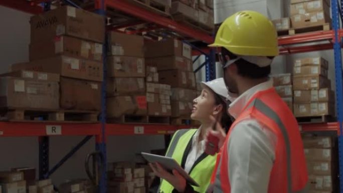 亚洲仓库工人男女在仓库与白色安全帽讨论使用数字平板电脑。仓库员工在分销仓库检查库存。