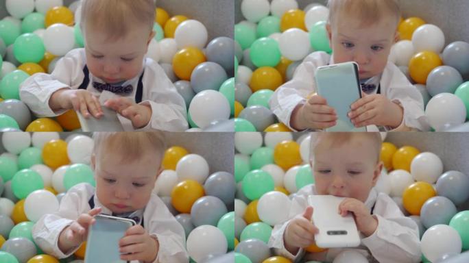 孩子在家里的球坑里玩智能手机。快乐的孩子躺在软球坑里，有五颜六色的球给幼儿，高加索男婴在家里玩得开心