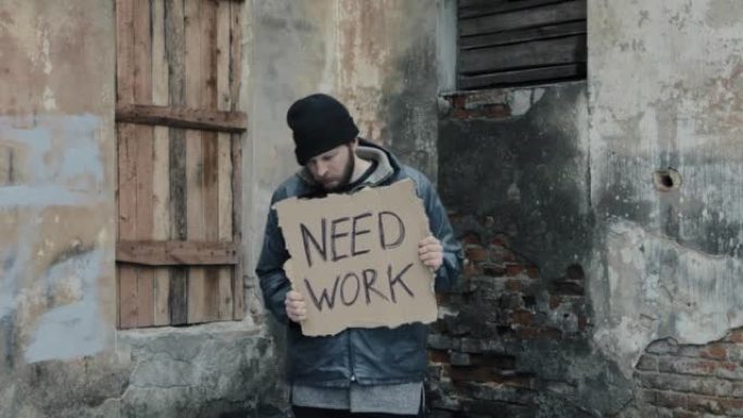 无家可归者拿着一张写着“需要工作”的纸板。难民正在找工作。穿着脏衣服的男流浪汉。生活在贫困线以下。大