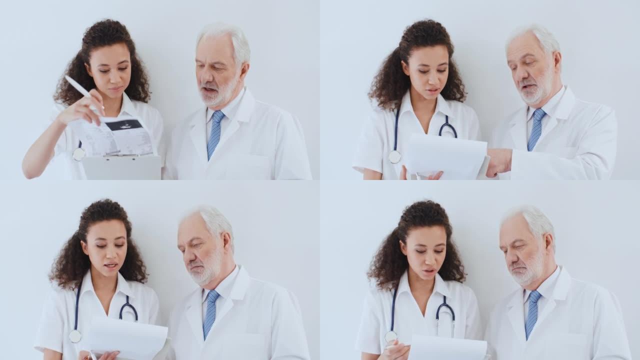 肖像两名医务人员老年白人男性和年轻卷发多种族女性，医生讨论医疗病例，在医院站在白墙边谈论健康报告