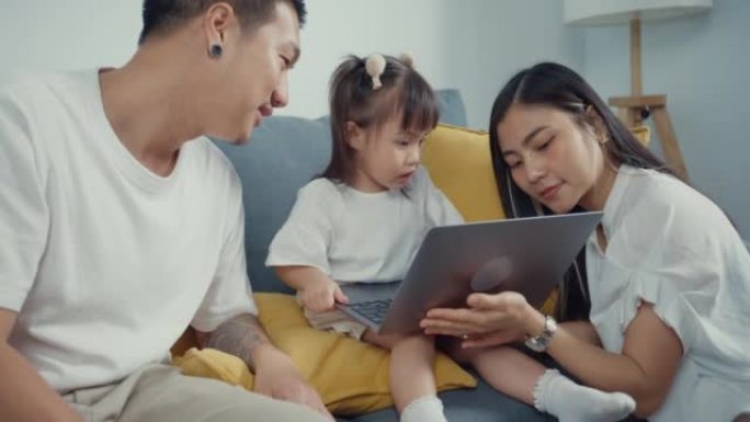 年轻的父母亚洲家庭坐在沙发上享受黄金时光快乐的时刻，和他们的孩子一起在家里的客厅用笔记本电脑看卡通电