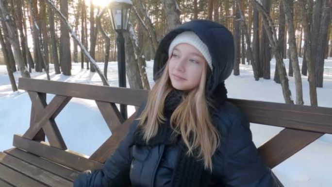 穿着黑色外套的体贴漂亮金发女孩坐在冬季白雪皑皑的公园的木凳上欣赏风景。背景上的灯笼。晴天。