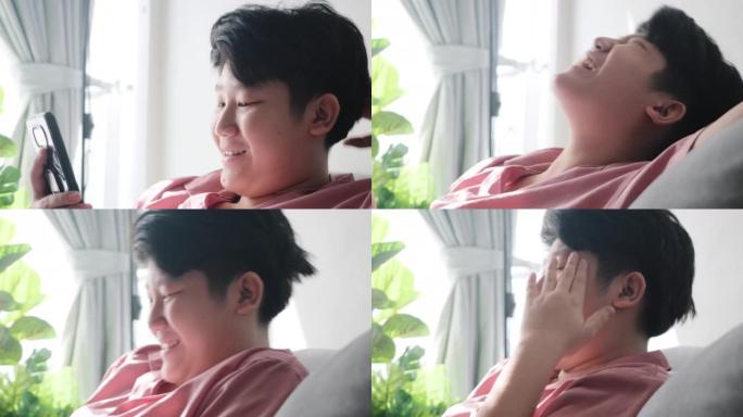 在家里用阳光照耀的沙发上看电影或病毒视频，在家里用生活方式概念笑亚洲男孩。