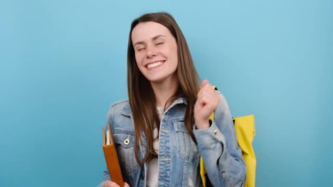 开朗的微笑女孩青少年学生拿着书跳舞休息，穿着牛仔夹克和黄色背包，在蓝色背景工作室中孤立地摆姿势。高中