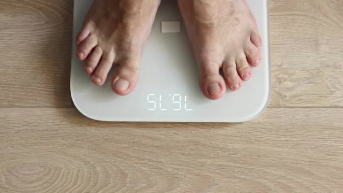 男性赤脚踩在白色数字地磅上-男人在家称自己: 特写俯视图。测量体重、技术、控制、健康和饮食概念