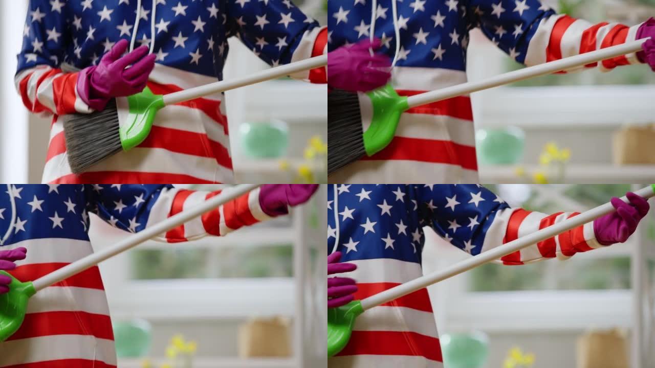 面目全非的家庭主妇模仿在扫帚打扫屋里弹吉他。穿着美国国旗连帽衫的开朗女士在室内客厅玩得开心。