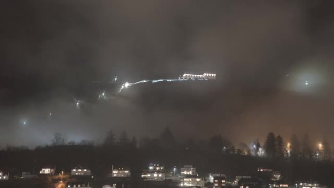 山顶上有弗洛伊巴宁缆车顶站照明建筑的浮山 -- 夜间时光倒流，雾气从挪威卑尔根山前穿过