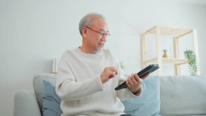 亚洲有吸引力的老年男子在家里的客厅使用平板电脑。年长的成熟祖父坐在沙发上戴着眼镜，在智能手机上聊天，