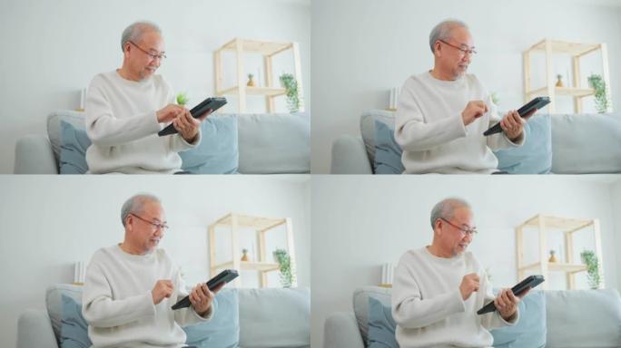 亚洲有吸引力的老年男子在家里的客厅使用平板电脑。年长的成熟祖父坐在沙发上戴着眼镜，在智能手机上聊天，