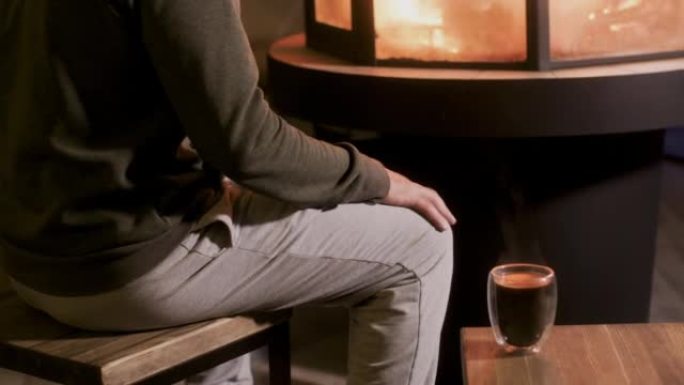 身穿灰色长裤的面目全非的男人坐在壁炉旁，拿着玻璃热茶，自己抚摸膝盖，在室内用火取暖。