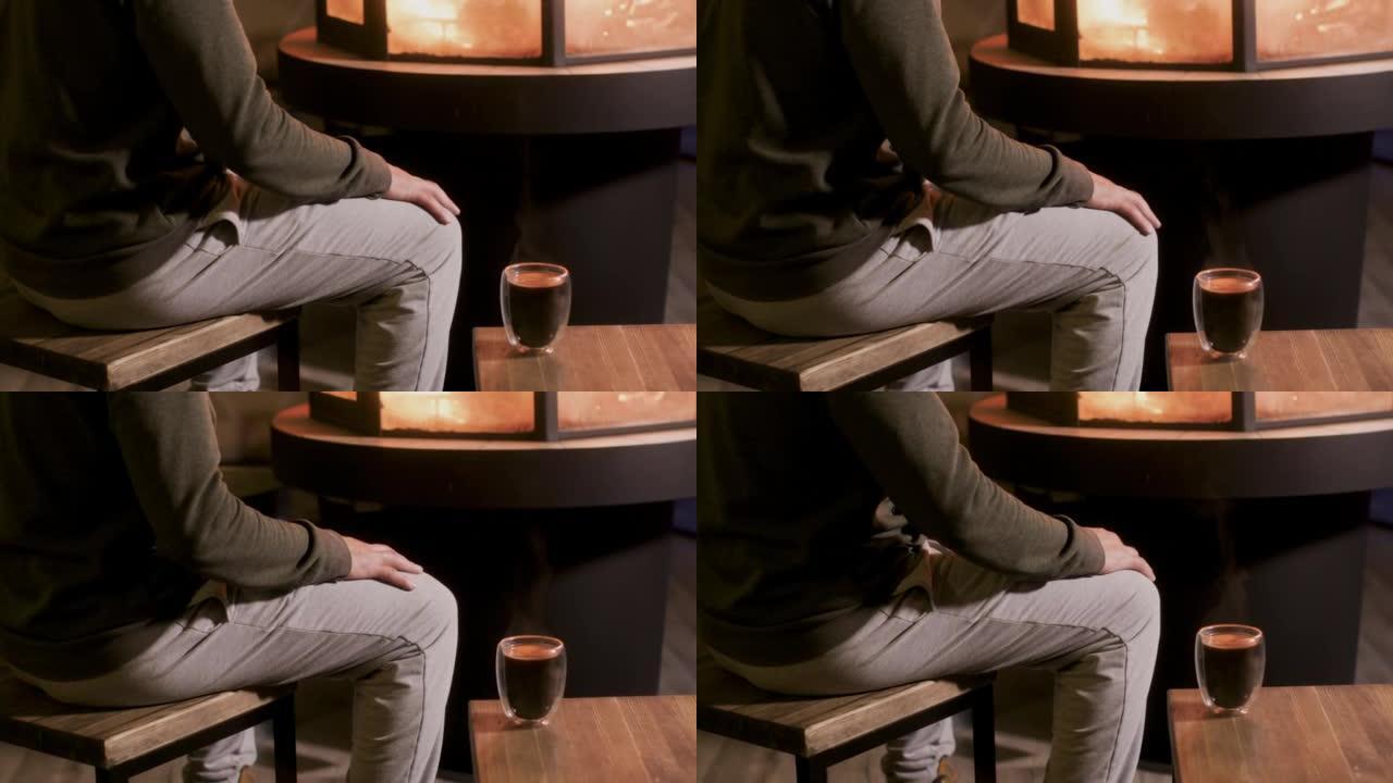 身穿灰色长裤的面目全非的男人坐在壁炉旁，拿着玻璃热茶，自己抚摸膝盖，在室内用火取暖。
