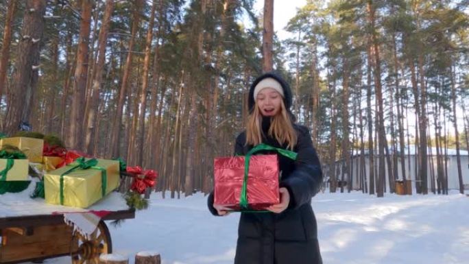 兴奋的少女在冬天的白雪皑皑的森林里抓住绿色丝带的红色礼品盒。背景上有圣诞彩盒的手推车。