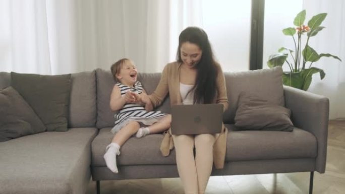 平静的年轻母亲坐在沙发上，在笔记本电脑上工作，小女孩拉着她的手，想玩。安静的妈妈在沙发上放松使用现代
