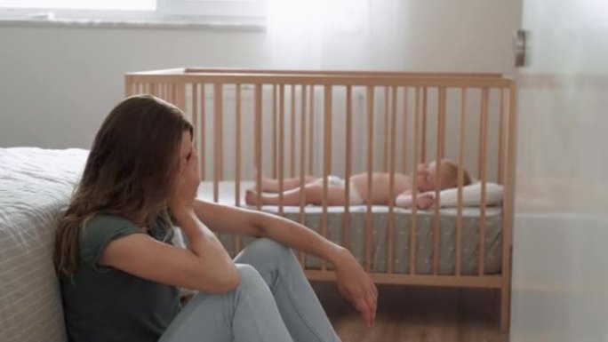 患有产后抑郁症的母亲坐在地板上，头跪在婴儿床旁边