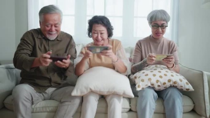 幸福亚洲退休朋友享受玩和比赛游戏智能手机在线沙发在客厅家庭室内背景，亚洲团体一起玩游戏家庭孤立的想法