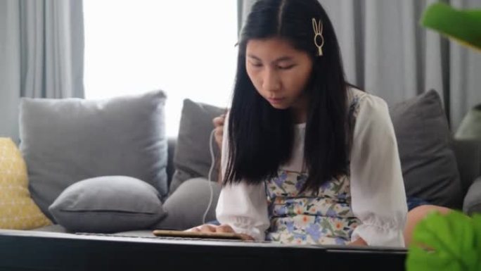 亚洲女孩在家里靠窗的沙发上通过移动应用学习弹奏键盘乐器，生活方式概念。