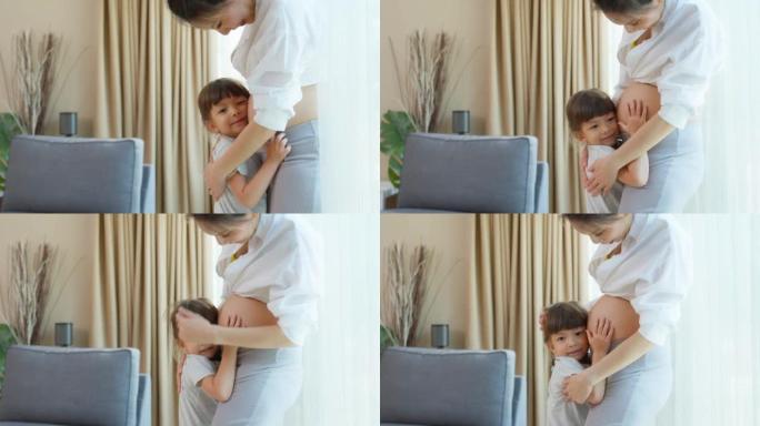亚洲小女儿拥抱抚摸美丽的怀孕妈妈肚子。迷人的怀孕女孩站在室内靠近客厅窗户的时候，用幸福和爱拥抱小女孩