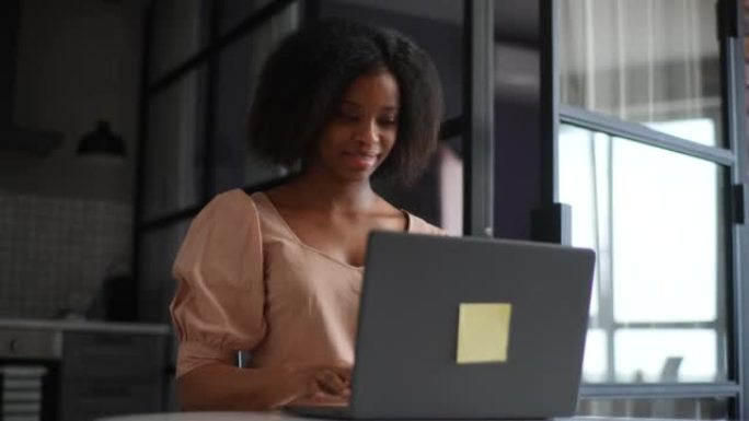 快乐的年轻非裔美国女商人坐在现代厨房的桌子上，在互联网上工作的笔记本电脑上打字。